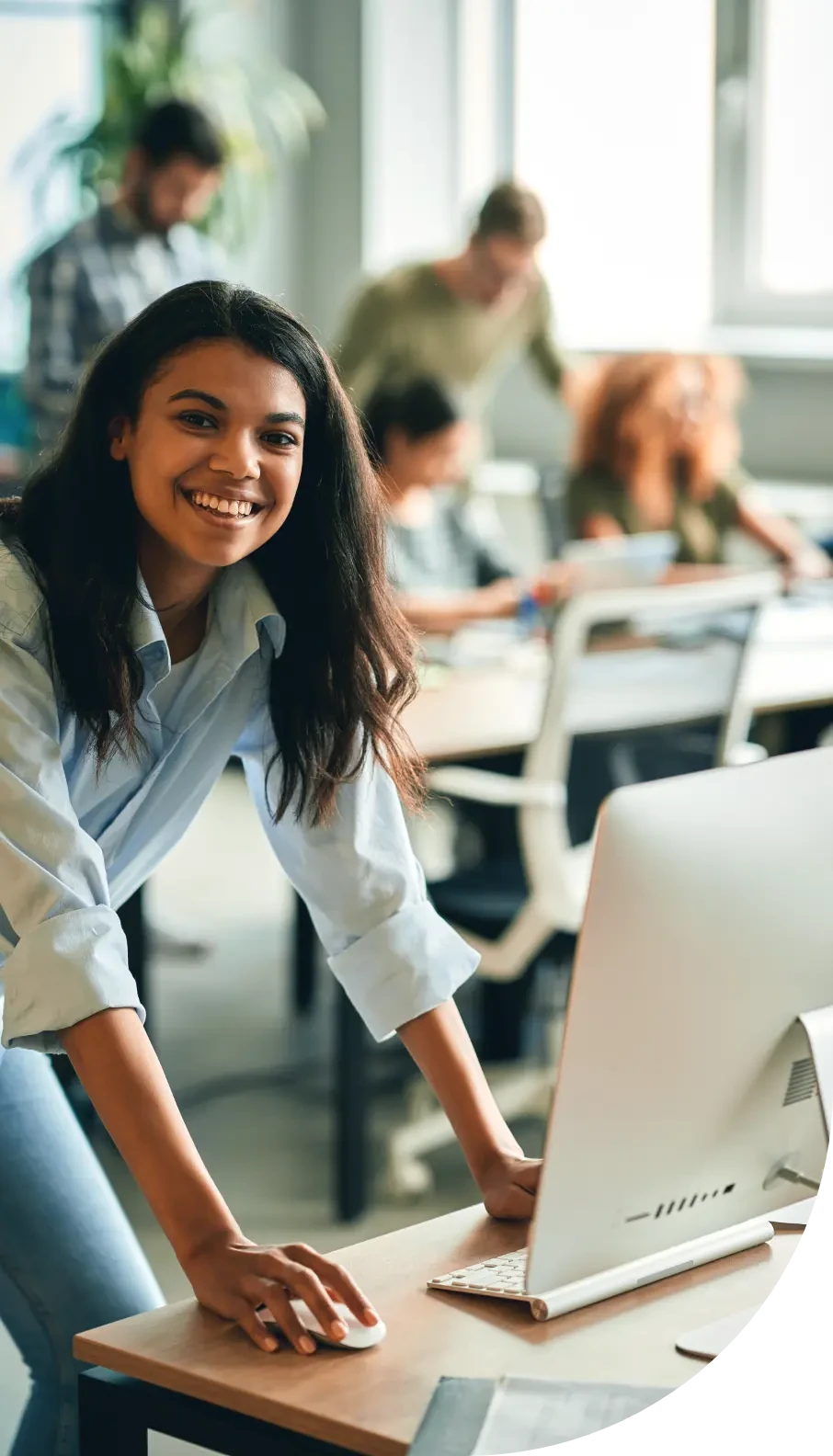A smiling woman leans over a desk in a bright, busy office.