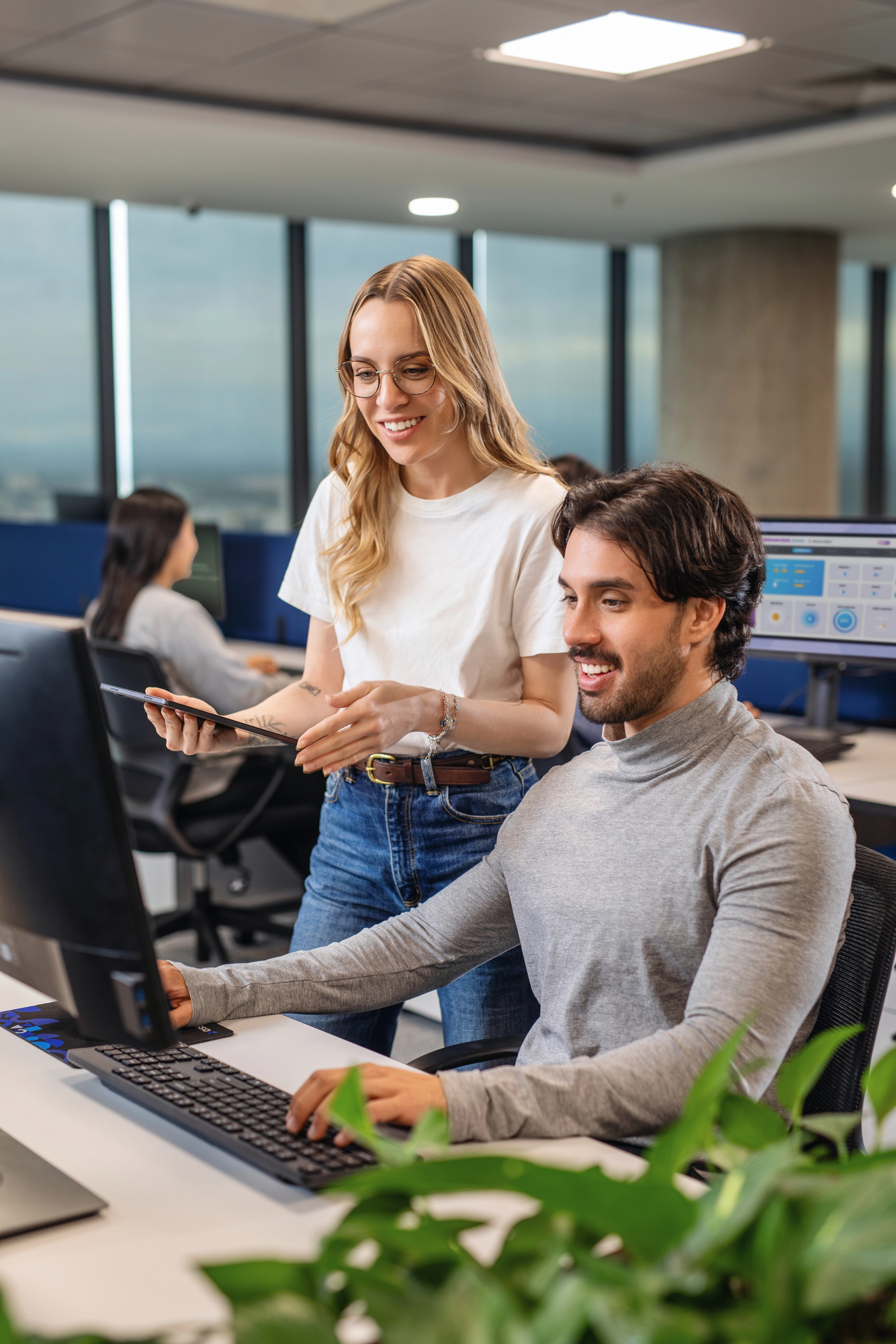 A smiling woman leans over a desk in a bright, busy office.