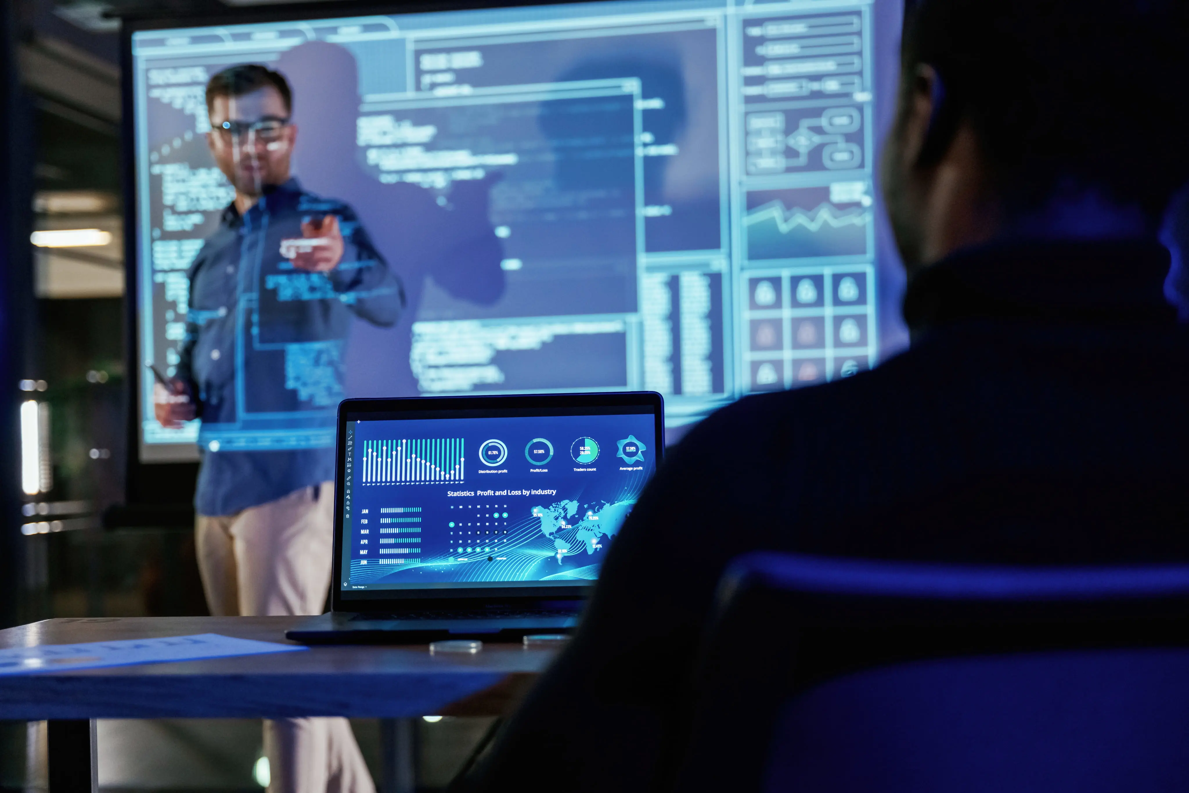 A person is presenting technical information on a large screen to someone working on a laptop in a high-tech setting.