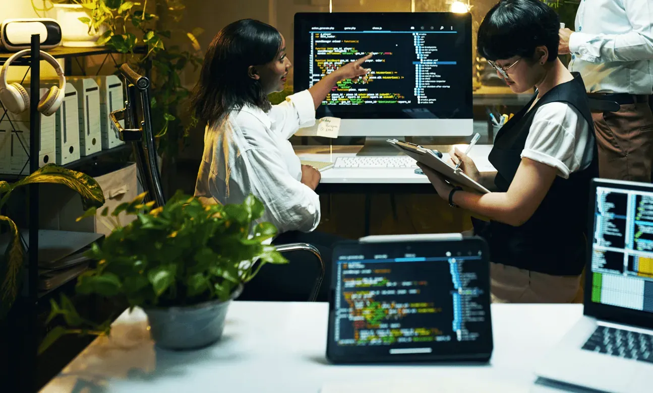 Three people work together in an office filled with multiple screens displaying code.