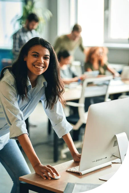 A smiling woman leans over a desk in a bright, busy office.