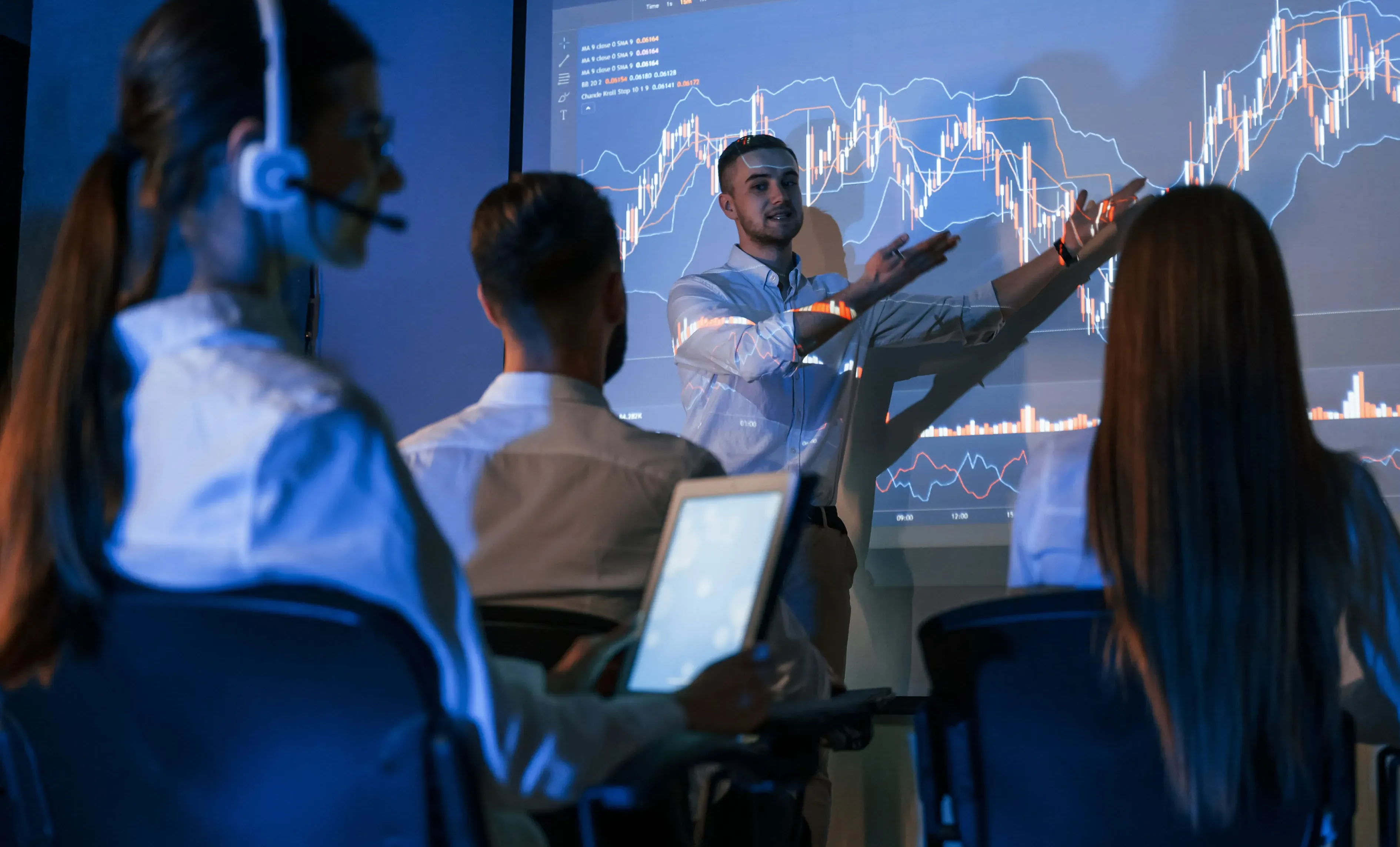 A presenter explains financial charts to a group of people in a darkened room.
